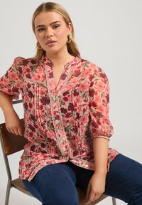 Blusa a maniche lunghe con bottoni, realizzata in un tessuto trasparente decorato con fiori rosa e rossi, con dettagli in pizzo e una vestibilità comoda, abbinata a jeans blu scuro.