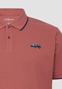 Rostfarbendes Polo-Shirt mit schwarzem Besatz am Kragen und den Ärmelenden, zwei Knöpfen und schwarzer Vintage-Auto-Stickerei auf der Brust.