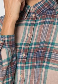 Camicia a quadri con bottoni in tonalità di teal, marrone e crema. Tessuto morbido con colletto e chiusura con bottoni. Maniche lunghe.