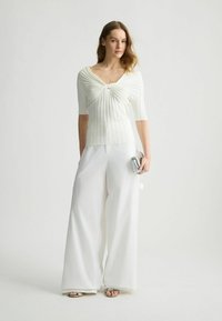 Top bianco plissettato con scollatura a V e maniche corte, abbinato a pantaloni bianchi larghi. Il modello tiene una piccola borsetta metallica, in piedi su uno sfondo neutro.