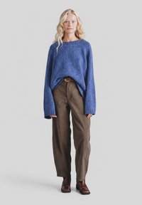 Kobieta o długich blond włosach, ubrana w luźny niebieski sweter z dzianiny, brązowe spodnie sztruksowe oraz ciemnobrązowe botki na kostkę, na jednolitym tle.