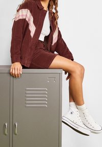 Jong persoon in een bordeaux corduroyjack en rok, zittend op een grijze metalen locker, met witte hoge Converse sneakers en witte sokken.
