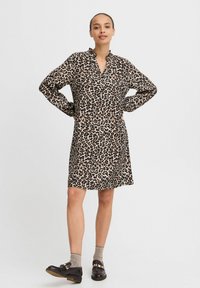 Leopardmönstrad klänning i beige och svart, med krage utan hals, långa ärmar och avslappnad passform. Matchad med svarta skor och grå strumpor.