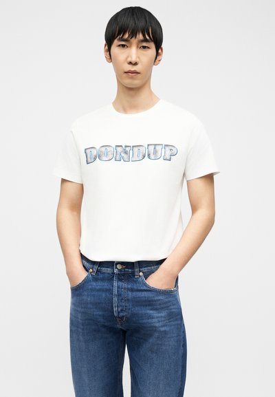 Νεαρός άνδρας με μαύρα μαλλιά, που φοράει λευκό μπλουζάκι με το κείμενο "DONDUP" και μπλε τζιν, στέκεται με τα χέρια στις τσέπες μπροστά σε λευκό φόντο.