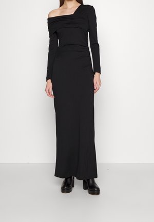 Femme portant une longue robe noire avec une épaule dénudée et une manche longue, associée à des bottines noires à plateforme.