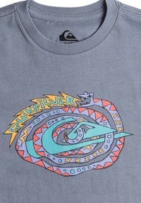 Šedé tričko s barevným, abstraktním designem spirálovitého hada a stylizovaným textem "Quiksilver" v žluté a tyrkysové na přední straně.