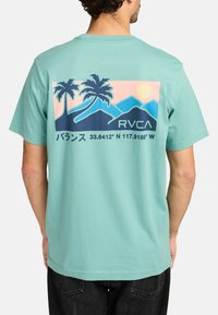 Mintgroene t-shirt met een graphic van palmbomen, bergen en een zon op de achterkant, met tekst en coördinaten in donker marineblauw.