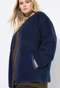 Junge Frau mit blonden Haaren, die eine dunkelblaue Fleecejacke mit Struktur und Taschen trägt, vor einem weißen Hintergrund stehend.