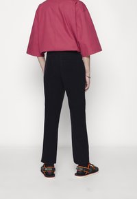 Volně padnoucí červená košile s krátkými rukávy, spojená s tmavými manšestrovými kalhotami a černými sandály s oranžovými akcenty. Pohled zezadu.