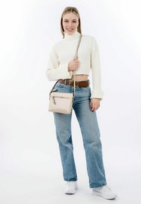 Beige Umhängetasche mit kontrastierenden Reißverschlussakzenten; weiche Textur. Modell trägt einen cremefarbenen gerippten Pullover, blaue Jeans und weiße Sneaker.