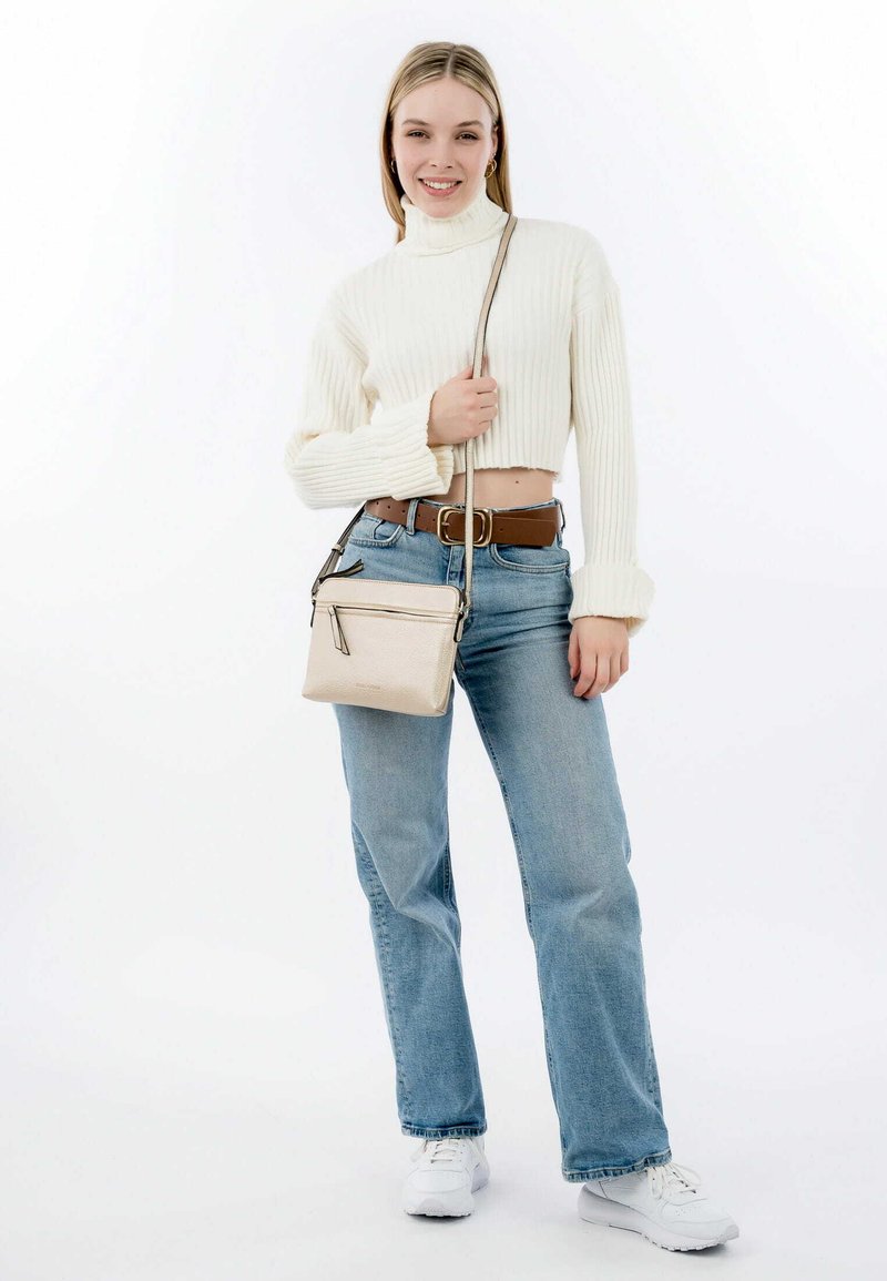 Beige crossbody bag with contrasting zipper accents; soft texture. Model wearing cream ribbed sweater, blue jeans, and white sneakers.