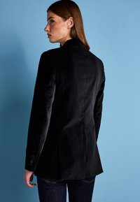 Blazer en velours noir avec une coupe ajustée, des revers crantés et des poignets à boutons. Texture lisse et design structuré.