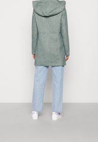 Giacca lunga, di un grigio-verde, con texture e cappuccio, abbinata a jeans azzurri larghi e scarpe bianche. La vista posteriore mostra la lunghezza del capo.
