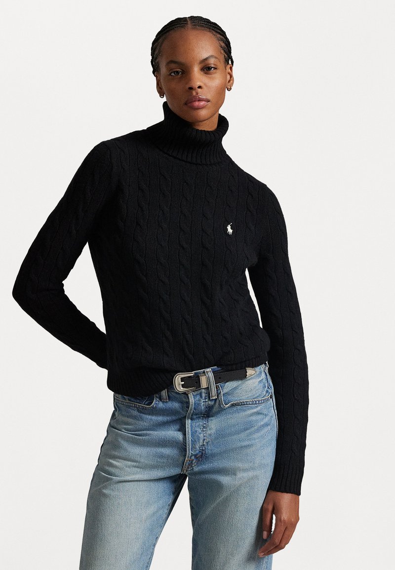 Svart kabelstickad tröja med hög polokrage, med en liten broderad logotyp på bröstet. Matchad med ljusblå jeans och ett svart bälte.
