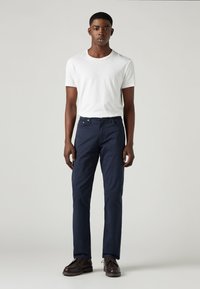 Marinblå chinos i bomullstwill, med rak skärning, två framfickor och bruna snörskor. Modellen bär en vit T-shirt.