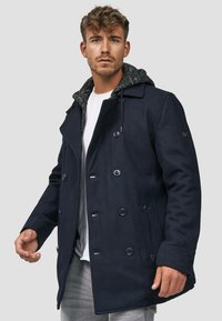 Marineblå dobbeltspent peacoat med teksturert grå hettefor; har store knapper og sidelommer, laget av ullblandingsstoff.