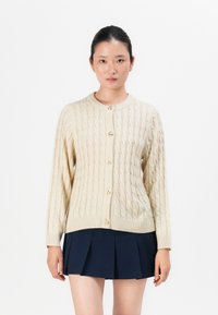 Ung kvinne med mørkt hår i en knute har på seg en beige kneppet cardigan med kabelstrikk og et marineblått plisserte skjørt mot en hvit bakgrunn.