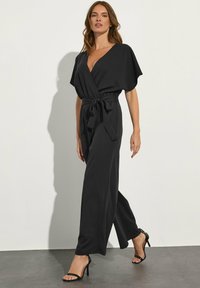 Tuta nera con un davanti avvolgente, maniche corte e vita legata. Presenta gambe ampie e una texture di tessuto liscia.