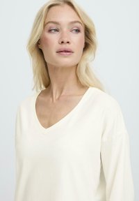 Tricou cu decolteu în V și mâneci lungi, de culoare crem, confecționat dintr-un material moale. Prezintă o croială lejeră și umeri puțin lăsați. Fără modele sau accente vizibile.