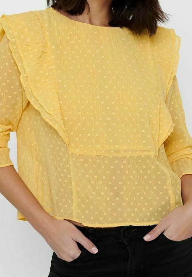 Gelbe durchsichtige Bluse mit gepunkteter Textur, Rüschendetails an den Ärmeln und vorne, mittellanger Schnitt, kombiniert mit schwarzen Hosen.