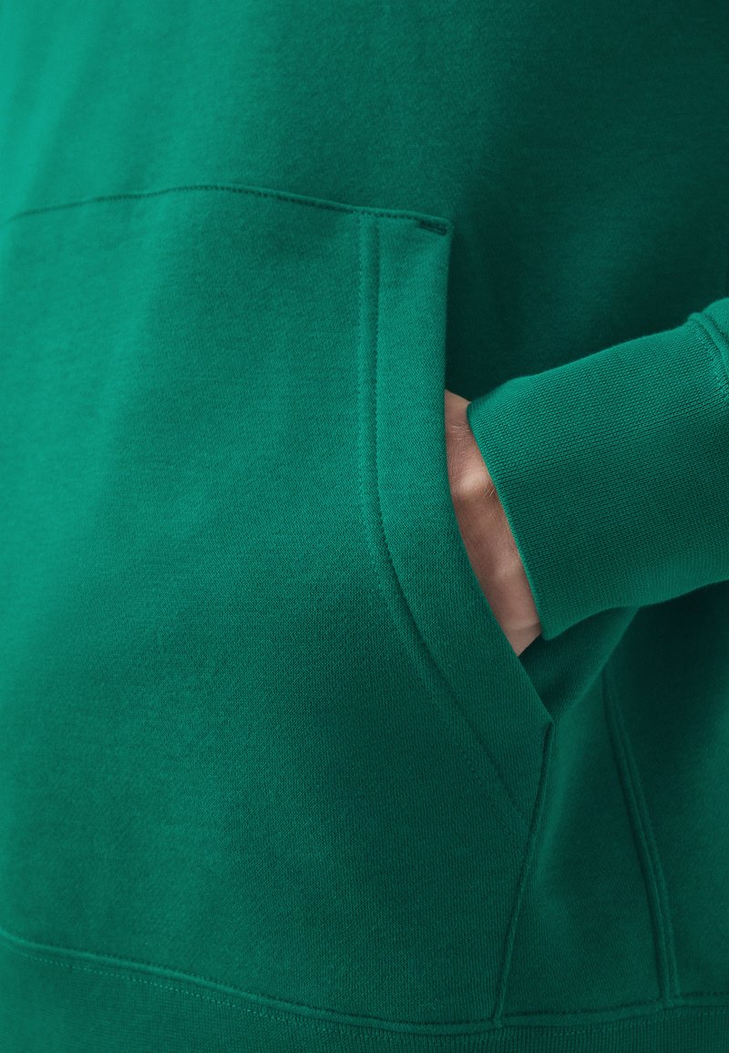 Sudadera verde con un bolsillo canguro, mangas acanaladas y una textura suave. Los detalles de costura a lo largo de las costuras añaden definición.