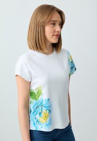 Weißes Baumwoll-T-Shirt mit floralem Design in Blau und Gelb auf der linken Seite, mit kurzen Ärmeln und Rundhalsausschnitt.