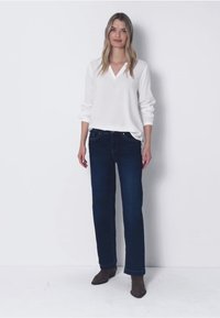 Donna in piedi, che indossa una maglietta bianca a maniche lunghe con scollo a V, jeans dritti blu scuro e stivaletti neri alla caviglia, su uno sfondo bianco semplice.