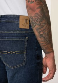 Jeans de mezclilla azul oscuro con un parche de piel etiquetado "JP 1880" en la cintura, que presentan costuras amarillas y un diseño sutil en el bolsillo trasero.