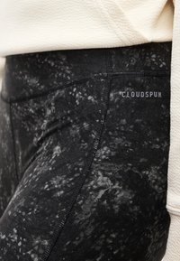 Κοντινό πλάνο σε μαύρα και γκρι κολάν με μαρμάρινο σχέδιο, με εμφανείς ραφές και λογότυπο "CLOUDSPUN" στο ύψος του γοφού, συνδυασμένα με ένα εκρού τοπ με υφή.