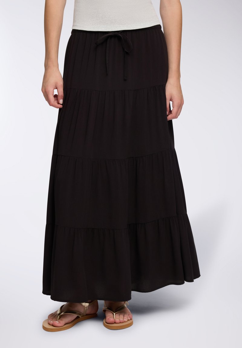 Kobieta w długiej, czarnej, warstwowej spódnicy z ściągaczem w talii oraz złotych płaskich sandałach, stojąca na jednolitym tle.