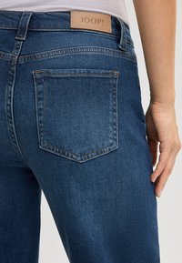 Jeans in denim di un blu profondo con una trama liscia. Presentano cinque tasche e una patch in pelle color nocciola etichettata "JOOP!" nella cintura.