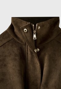 Gros plan du col d'une veste en daim marron avec une fermeture éclair argentée partiellement ouverte et deux boutons-pression argentés fermés à l'avant.