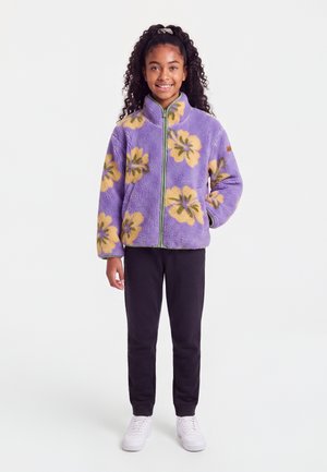 Fille souriante aux cheveux bouclés portant une veste polaire violette avec des fleurs jaunes, un pantalon noir, des baskets blanches, debout sur un fond uni.