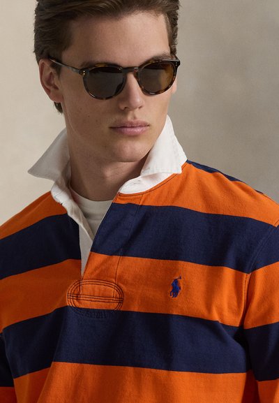 Oranges und marineblau gestreiftes Rugbyshirt mit weißem Kragen, halbknöpfbarem Verschluss und kleinem gesticktem Logo. Sonnenbrille mit rundem Rahmen.