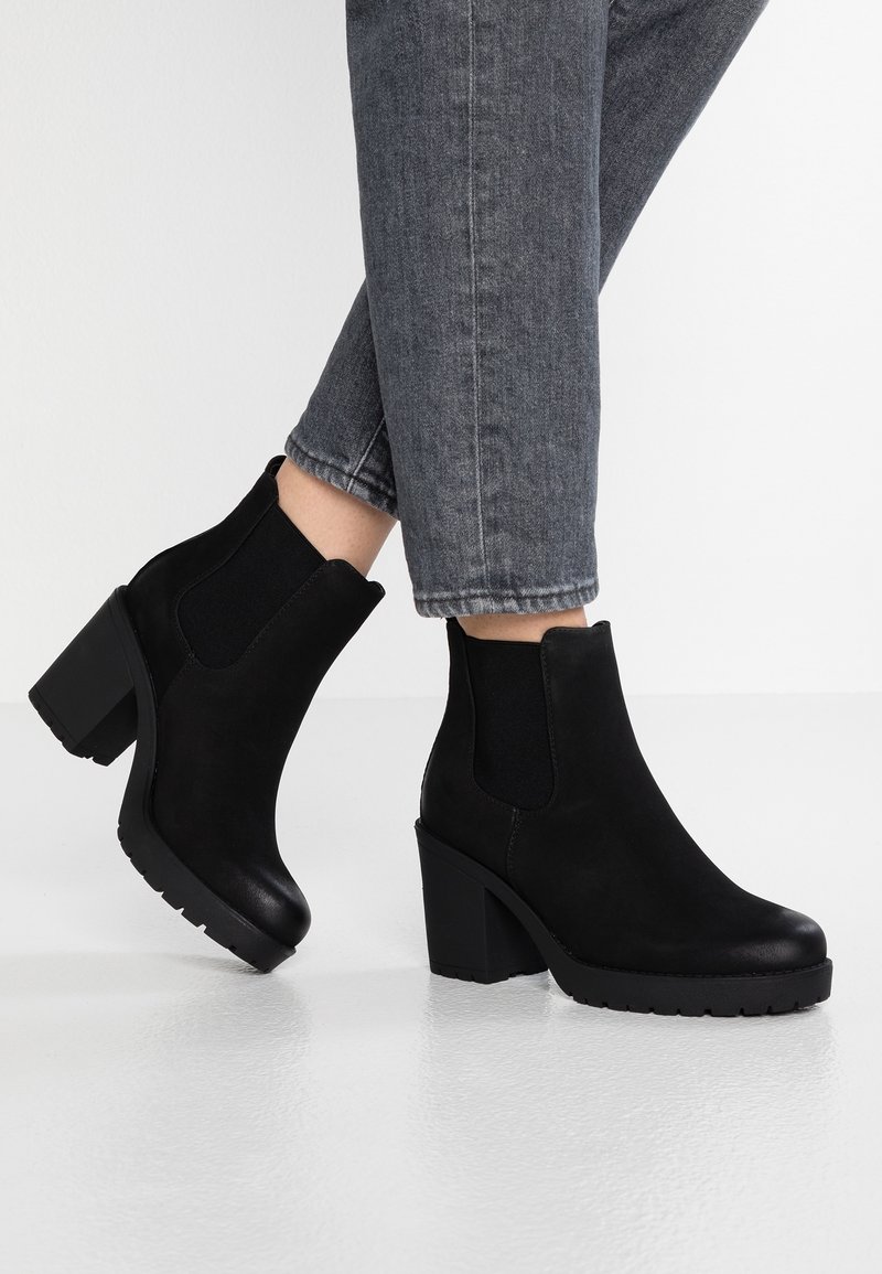 Bottines en daim noir à talons blocs portées avec un jean gris raccourci, présentées sur un fond de sol blanc brillant.