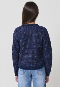 Maglione lavorato a maglia blu navy con un motivo testurizzato, colletto tondo e polsini a coste. Indossato con jeans azzurri chiari caratterizzati da cuciture decorative.