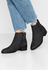 Botas negras hasta el tobillo con un acabado mate suave, cremallera lateral y detalle trenzado en el lado exterior. Tacón cuadrado. Llevadas con jeans azul desgastados.