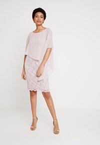 Robe rose pâle, longueur genou avec un overlay en dentelle et un ourlet festonné, associée à une cape semi-transparente. Le mannequin porte des talons dorés.