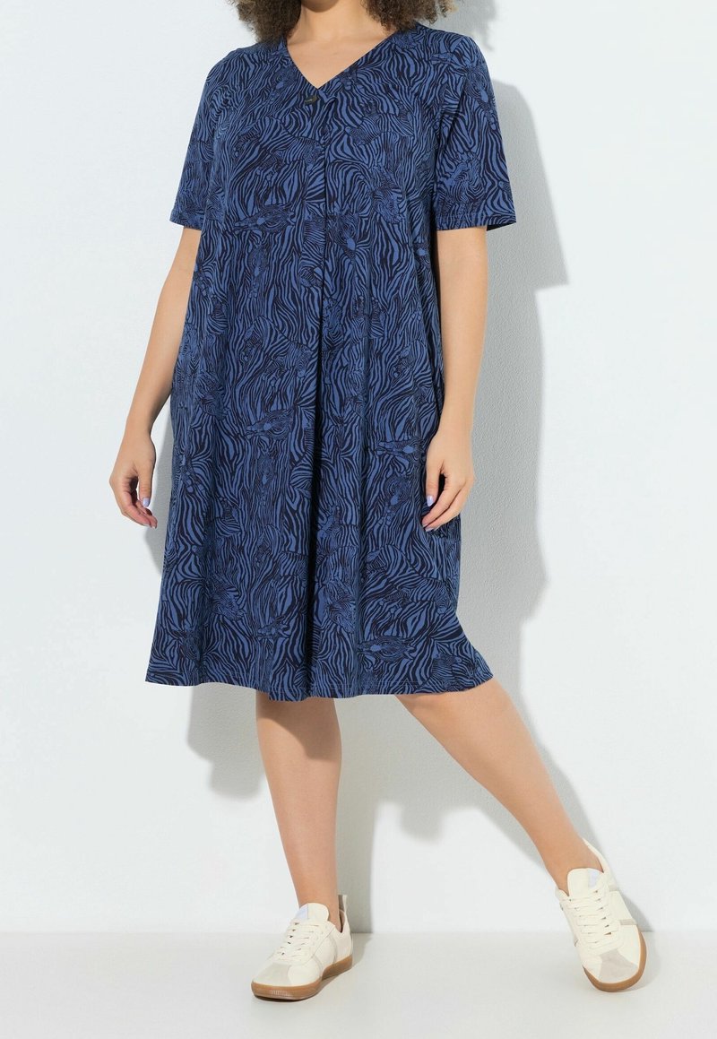 Femme portant une robe bleu marine au genou, manches courtes, motifs abstraits noirs, et des baskets blanches, debout contre un mur blanc uni.
