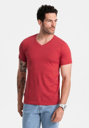 Homme avec des cheveux bouclés courts, bras gauche tatoué, T-shirt rouge à col en V, jeans bleus et montre blanche au poignet, regardant vers sa droite.