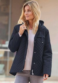 Gequilted zwarte jas met een ritssluiting en drukknopen, capuchon en zijzakken. Het heeft een zachte textuur en is gecombineerd met een lichtgekleurde trui.