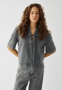 Top grigio in denim a maniche corte con lacci davanti, dettagli a occhiello e vestibilità rilassata. Il tessuto ha un aspetto morbido e strutturato.