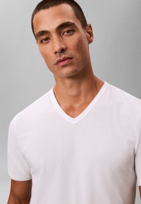 Jonge man met kort haar die een effen wit T-shirt met V-hals draagt tegen een grijze achtergrond.
