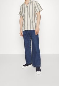 Camisa de manga corta con botones en crema con rayas verticales en azul marino, combinada con vaqueros de pierna ancha en azul y zapatillas azul marino.