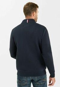 Felpa blu navy con collo a zip, caratterizzata da una striscia rossa, bianca e blu sul retro. Tessuto morbido e testurizzato; design aderente.