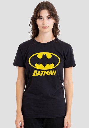 Young woman with brown hair wearing a black t-shirt featuring a large yellow Batman logo and text on the front against a plain background.