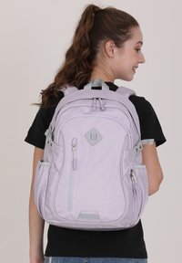 Sac à dos violet clair en tissu texturé, comportant plusieurs compartiments à fermeture éclair, des bretelles rembourrées et une poignée supérieure pour un transport facile.
