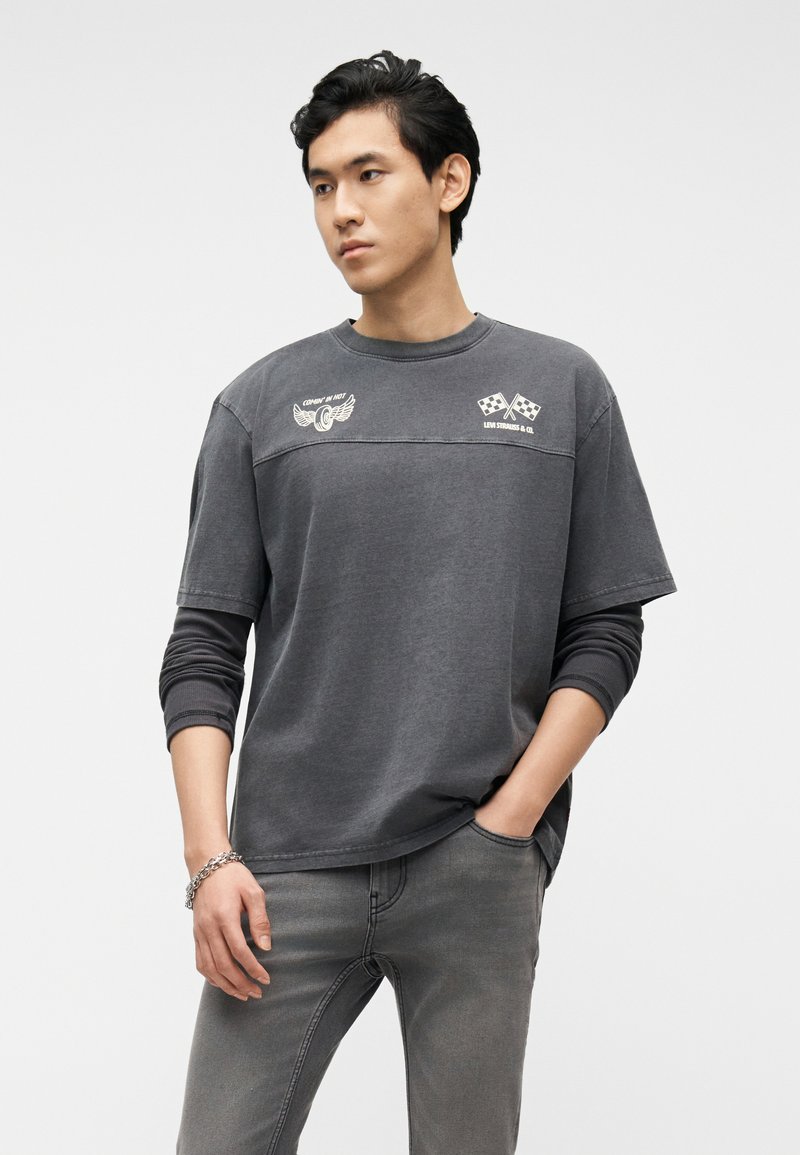 Young man with short black hair wearing layered dark gray shirts with printed logos and gray jeans, standing with one hand in pocket.