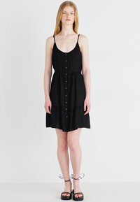 Rochie de soare neagră, cu butoane, cu un corset ajustat, fustă stratificată și bretele subțiri ajustabile. Pur portantă cu sandale cu platformă negre.