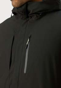 Giacca nera impermeabile con colletto alto, tasca sul petto con zip e dettagli grigi nella cerniera. Texture liscia con design minimalista.